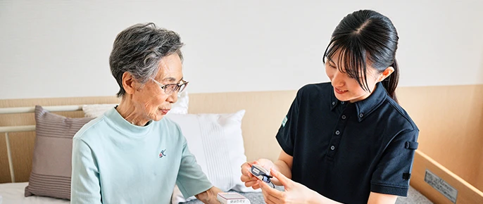 居室でスタッフが入居者に対応している様子。血圧測定や健康管理の場面。
